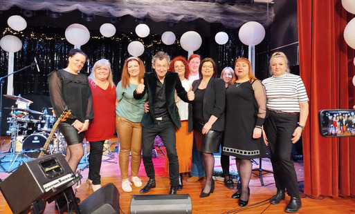 Grupa kobiet stoi na scenie razem z artystą po koncercie. W tle widać instrumenty muzyczne, dekoracyjne lampiony oraz sceniczne oświetlenie. Wszyscy uśmiechają się do zdjęcia, a mężczyzna stojący w środku pokazuje gest uniesionych kciuków. Scena udekorowa